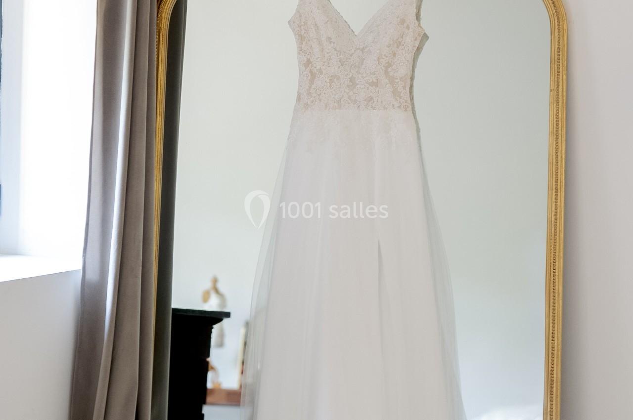 Robe de mariée blanche suspendue à un miroir doré dans une pièce lumineuse avec des rideaux gris.