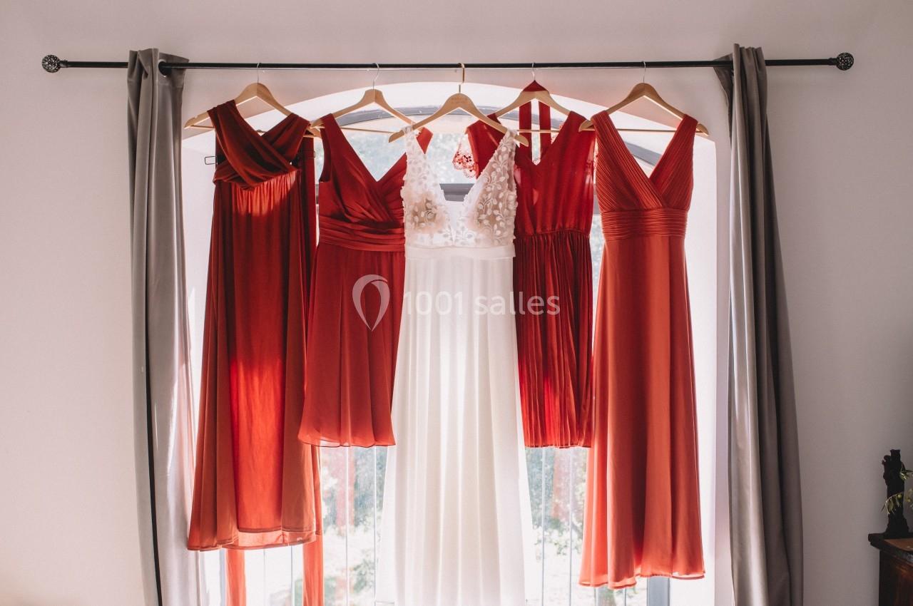 Quatre robes rouges et une robe blanche suspendues sur des cintres devant une fenêtre lumineuse.