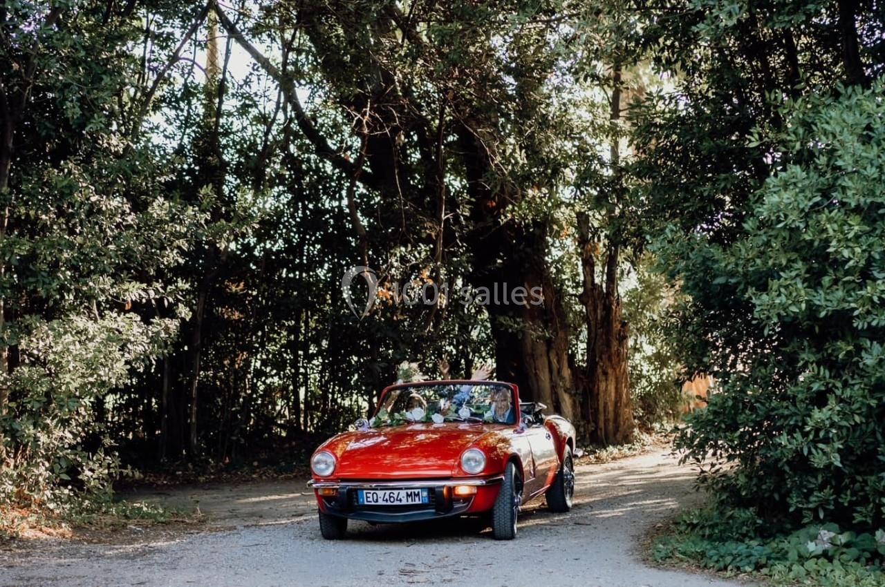 Voiture décapotable rouge garée sur un chemin de terre entouré d'arbres et de végétation dense.