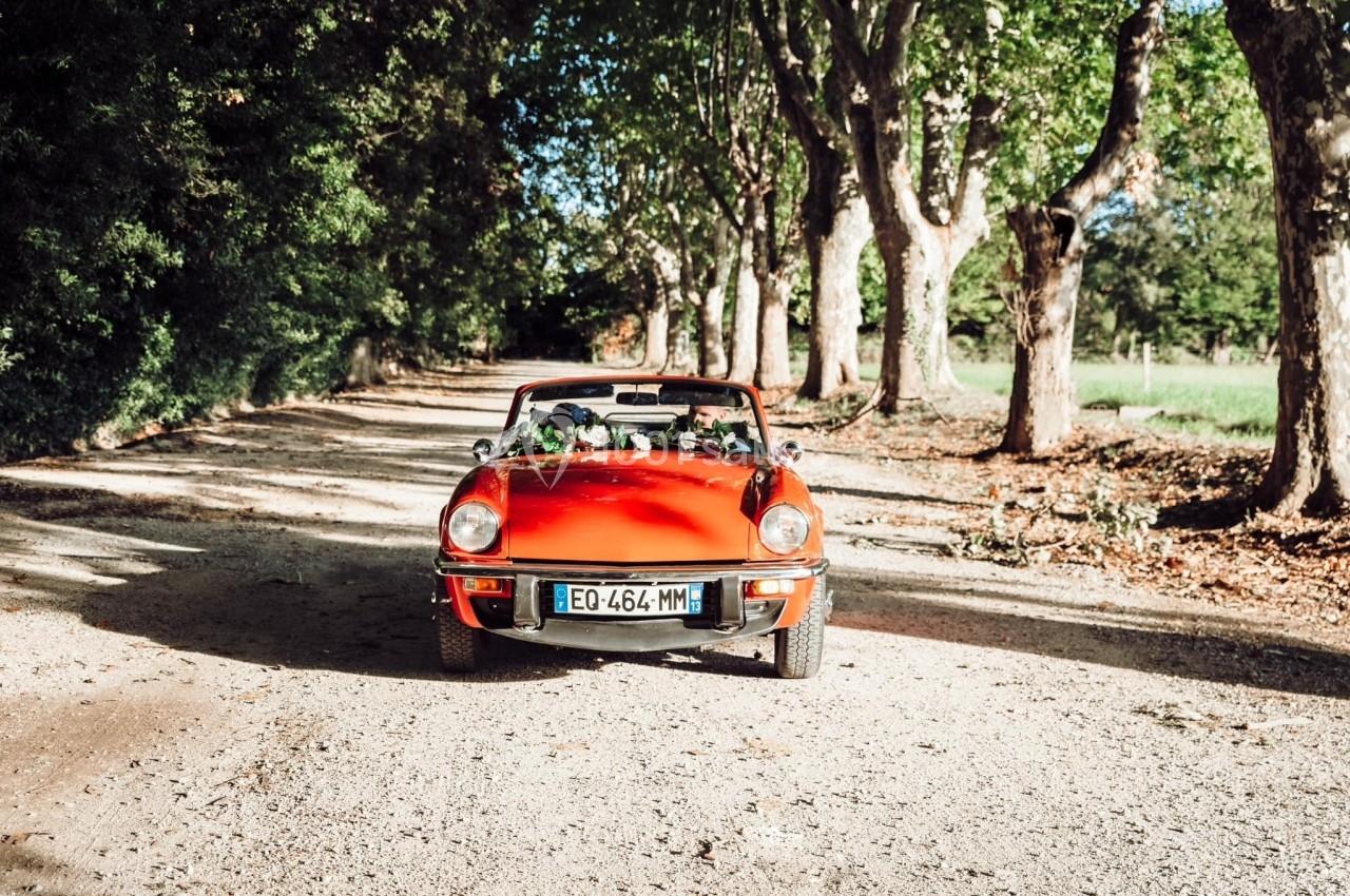 Voiture décapotable rouge circulant sur une route de campagne bordée d'arbres sous un ciel ensoleillé.