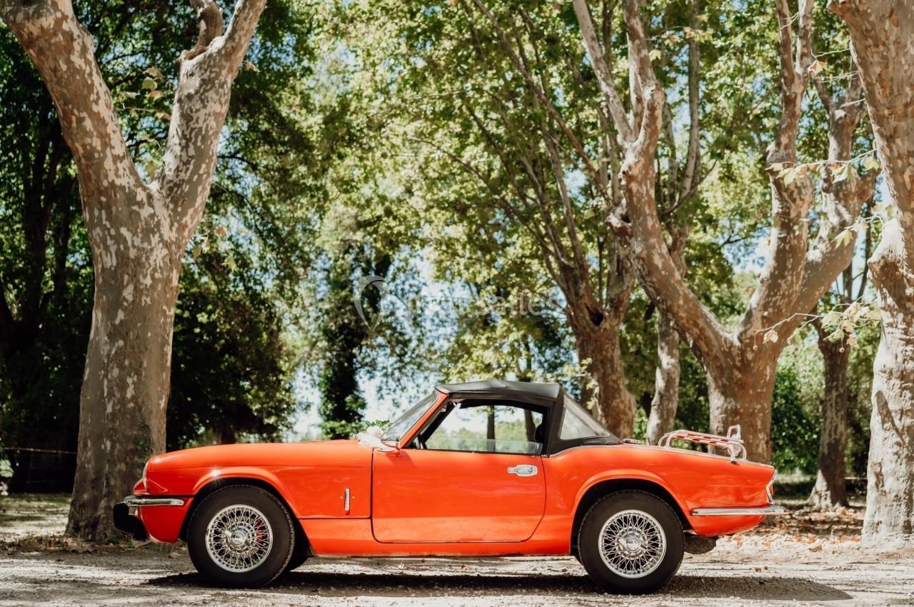 Voiture ancienne rouge décapotable garée sous des arbres dans un environnement ensoleillé.