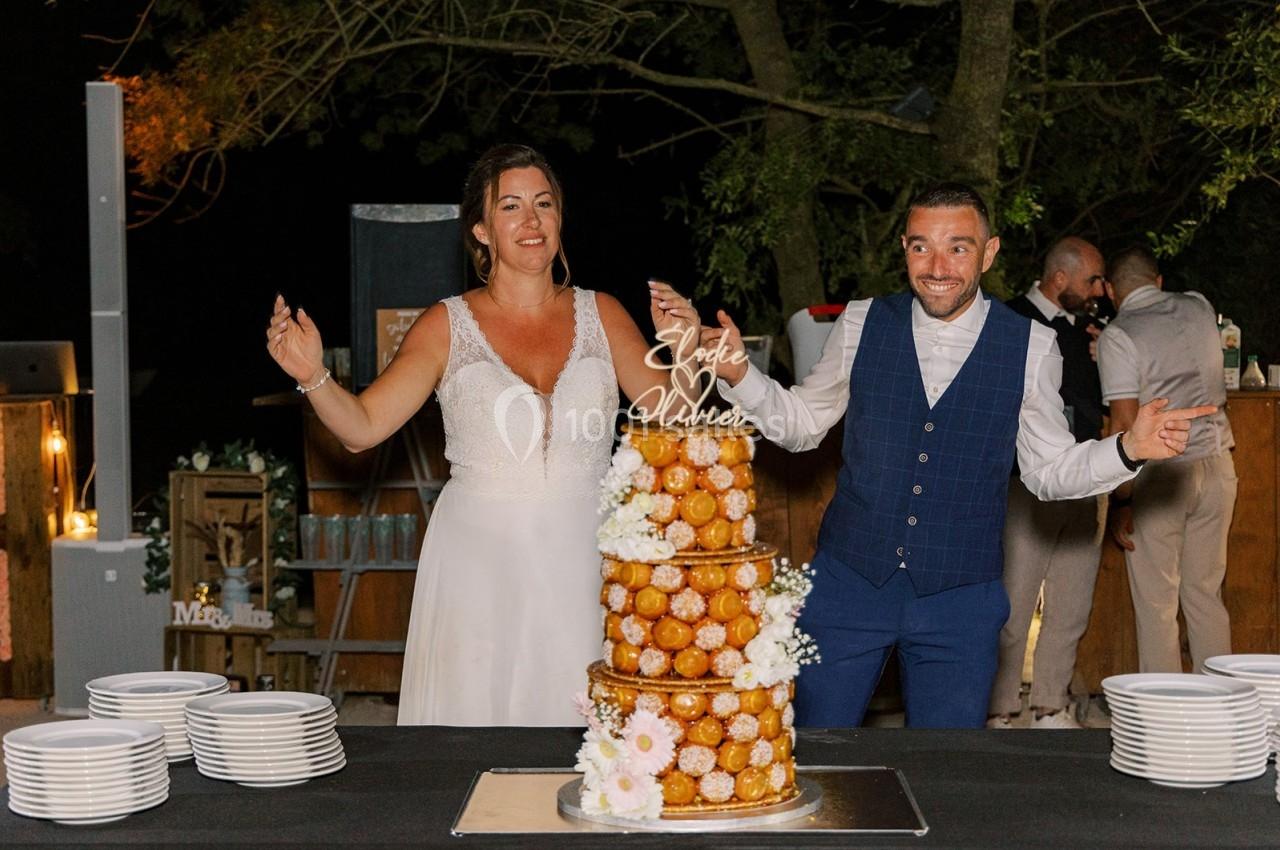 Un couple souriant pose devant une pièce montée de choux décorée de fleurs lors d'une réception en extérieur.