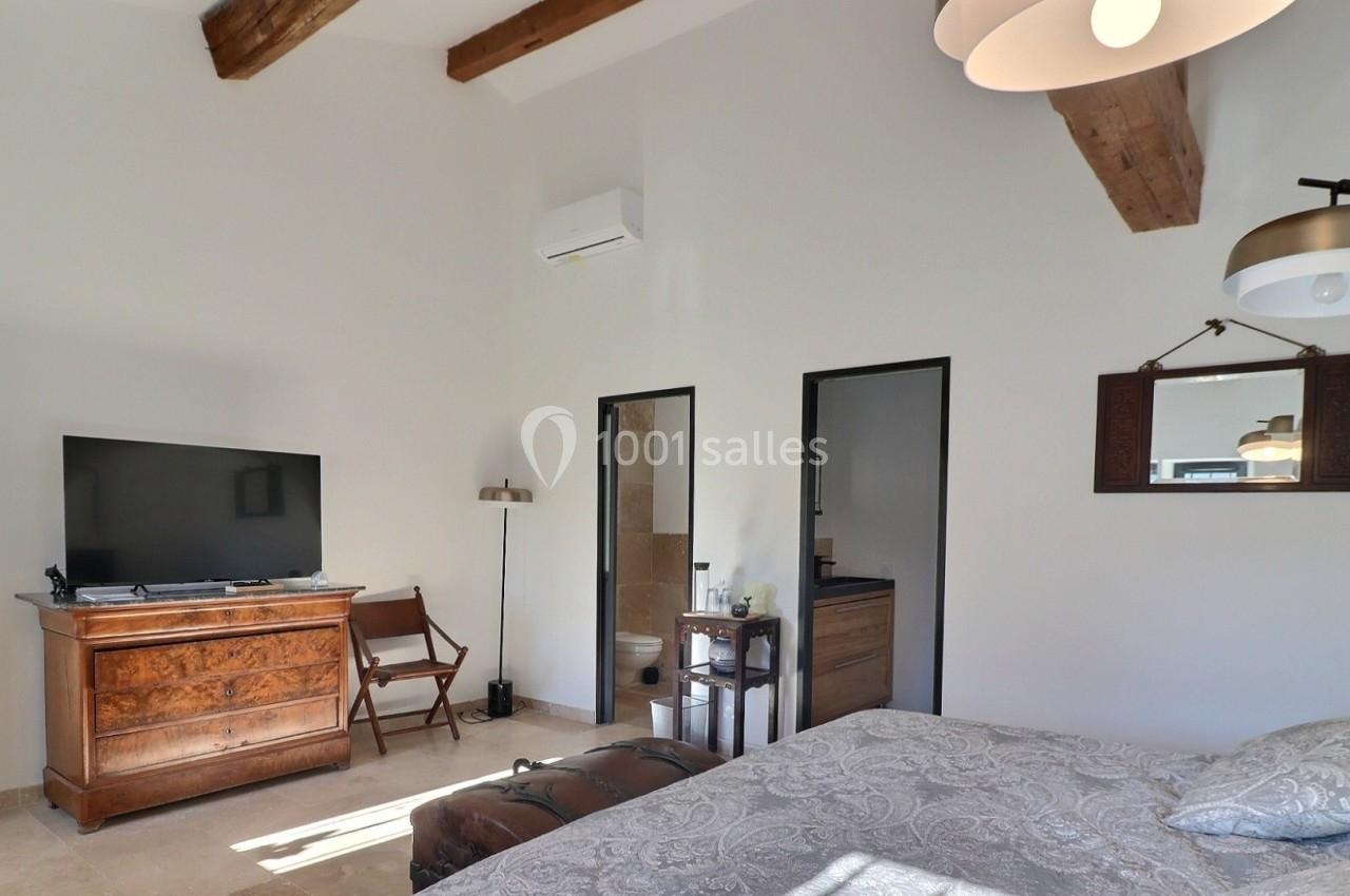 Chambre lumineuse avec lit double, poutres apparentes, téléviseur, commode en bois et salle de bain attenante.