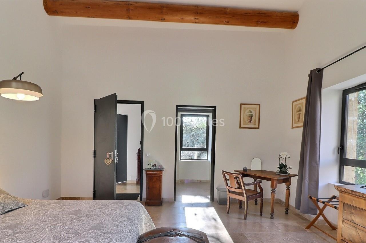 Chambre lumineuse avec lit double, bureau en bois, télévision, poutre apparente et vue sur l'extérieur par deux fenêtres.