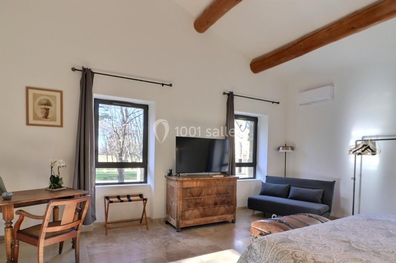 Chambre lumineuse avec plafond en poutres apparentes, bureau en bois, canapé, lit, télévision et fenêtres donnant sur un…