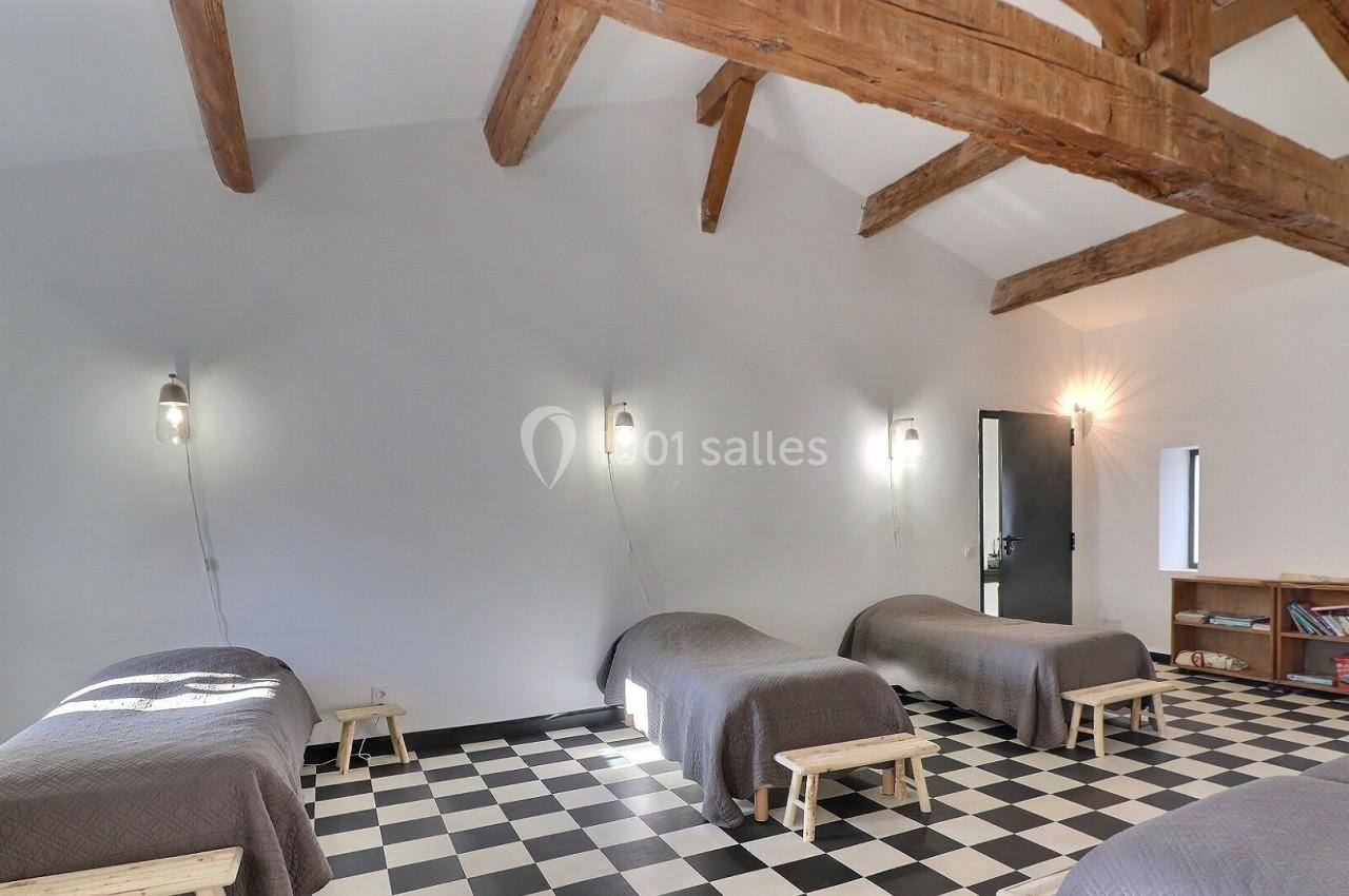 Quatre lits simples alignés dans une pièce lumineuse avec plafond en poutres apparentes et sol en damier noir et blanc.