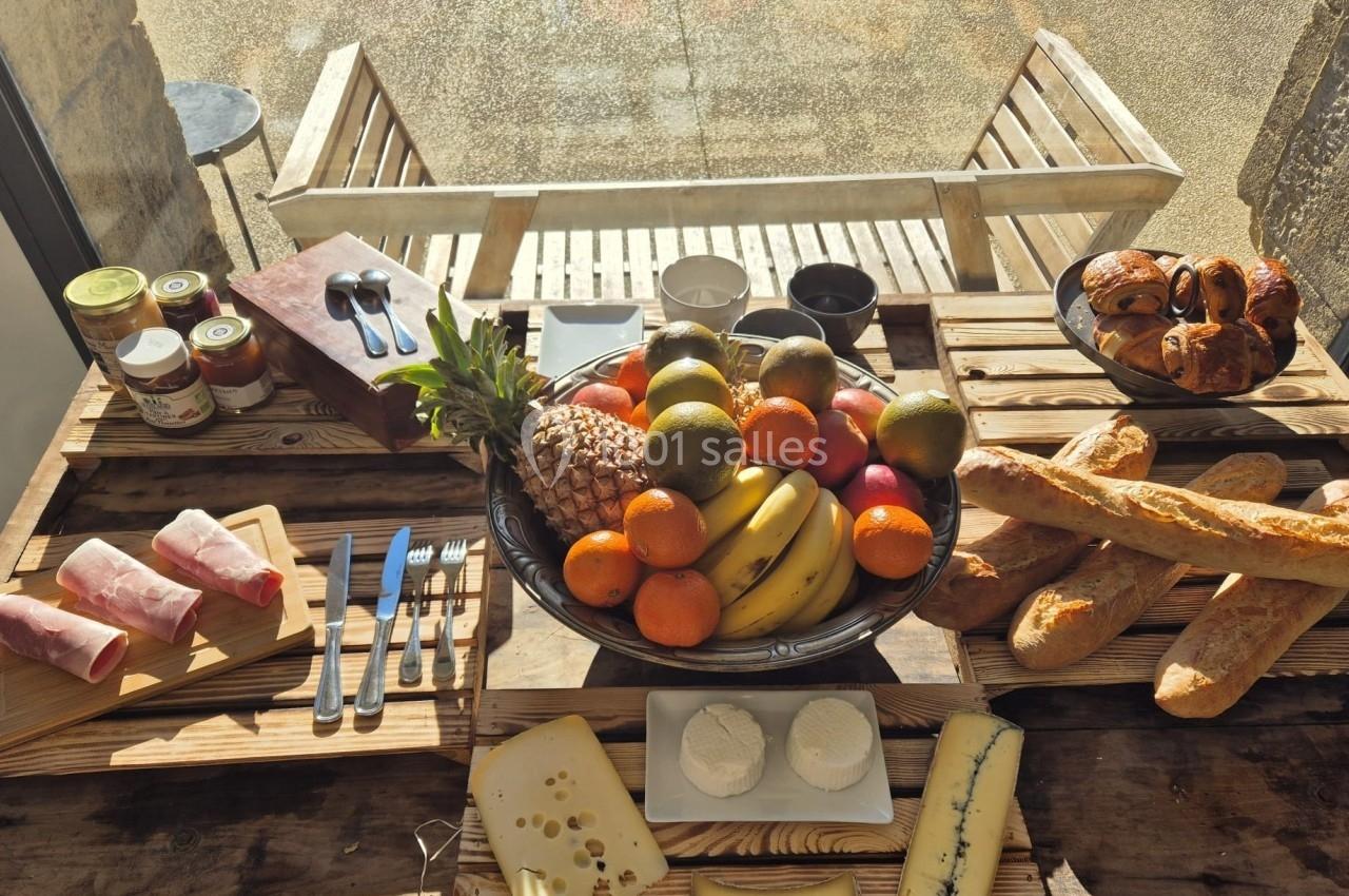 Petit-déjeuner avec fruits, pains, fromages, charcuterie et confitures disposés sur une table en bois près d'une fenêtre.