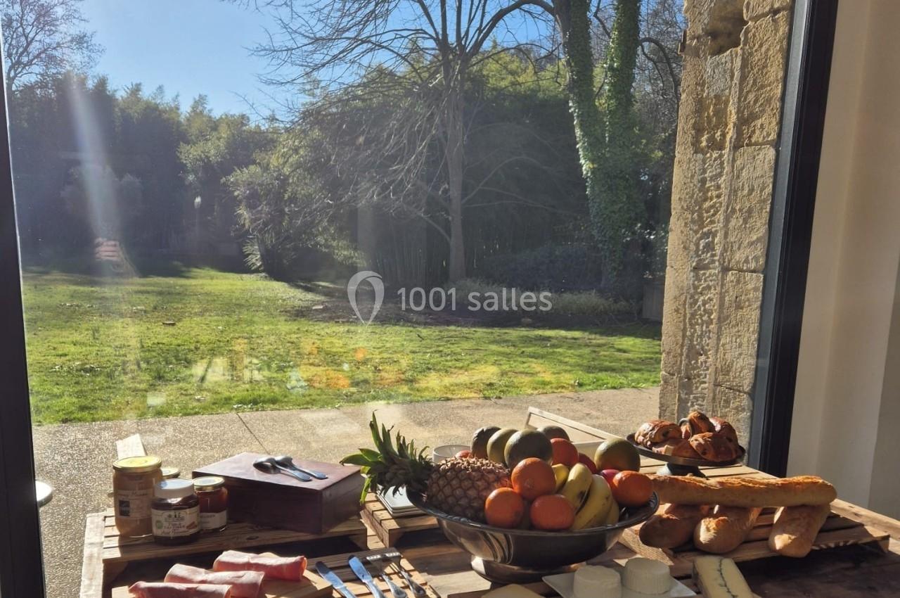 Table en bois avec charcuterie, fromages, fruits et pain, placée devant une baie vitrée donnant sur un jardin ensoleillé.