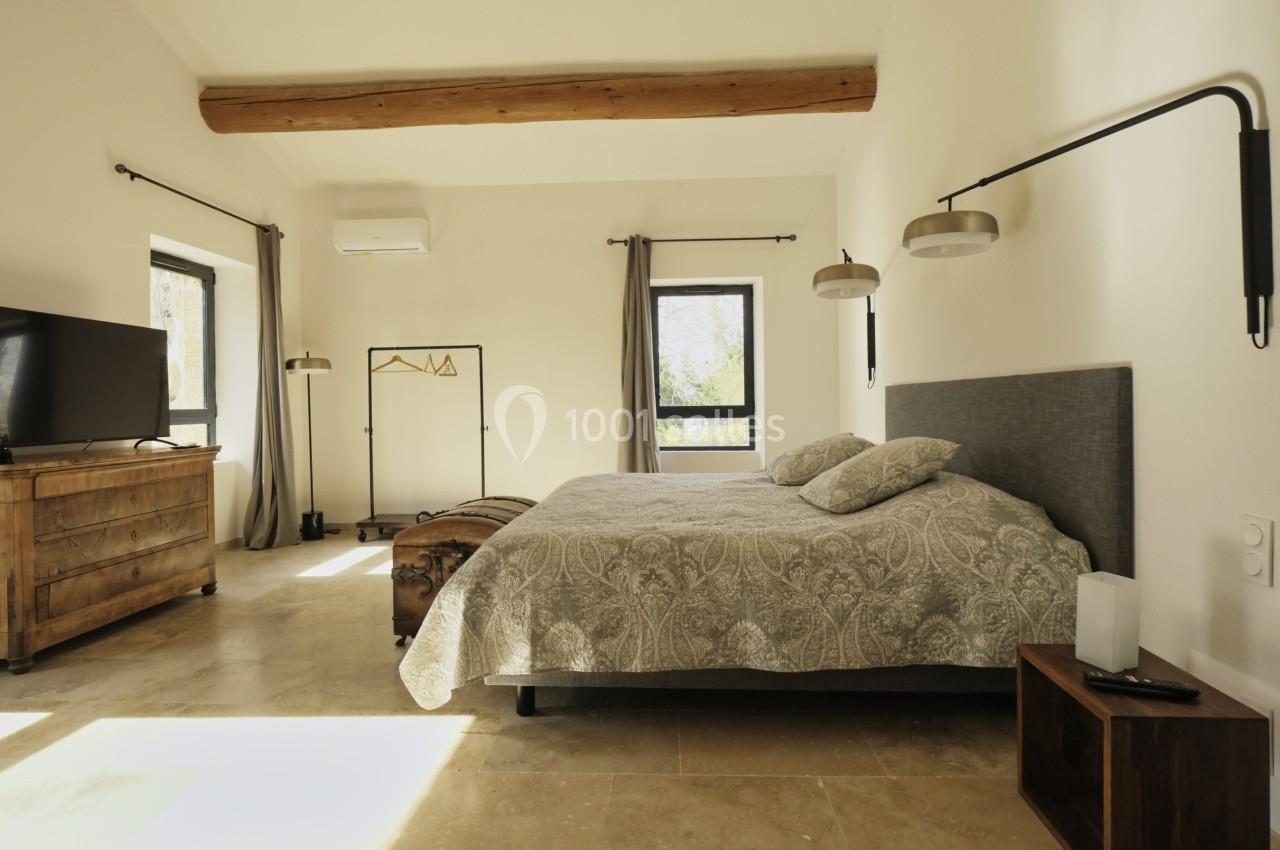 Chambre lumineuse avec un lit double, mobilier en bois, murs clairs, poutre apparente et grandes fenêtres.