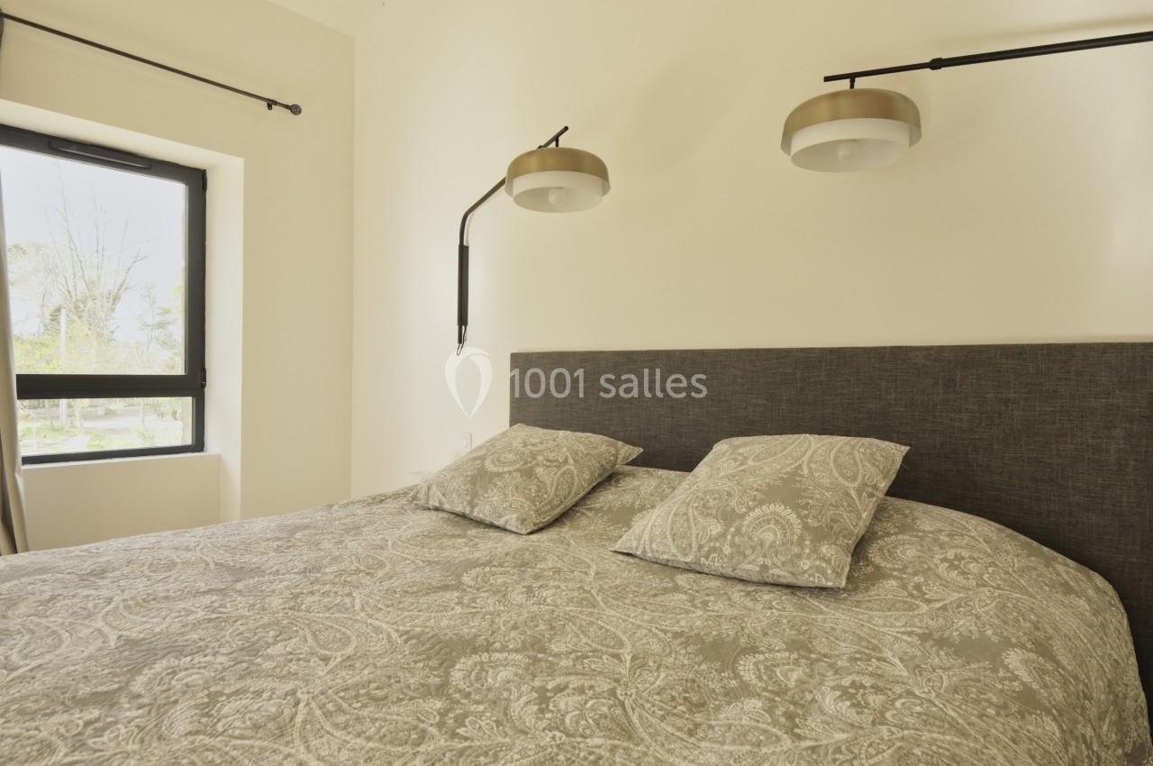 Lit double avec tête de lit grise, deux coussins assortis et luminaires muraux dans une chambre lumineuse.