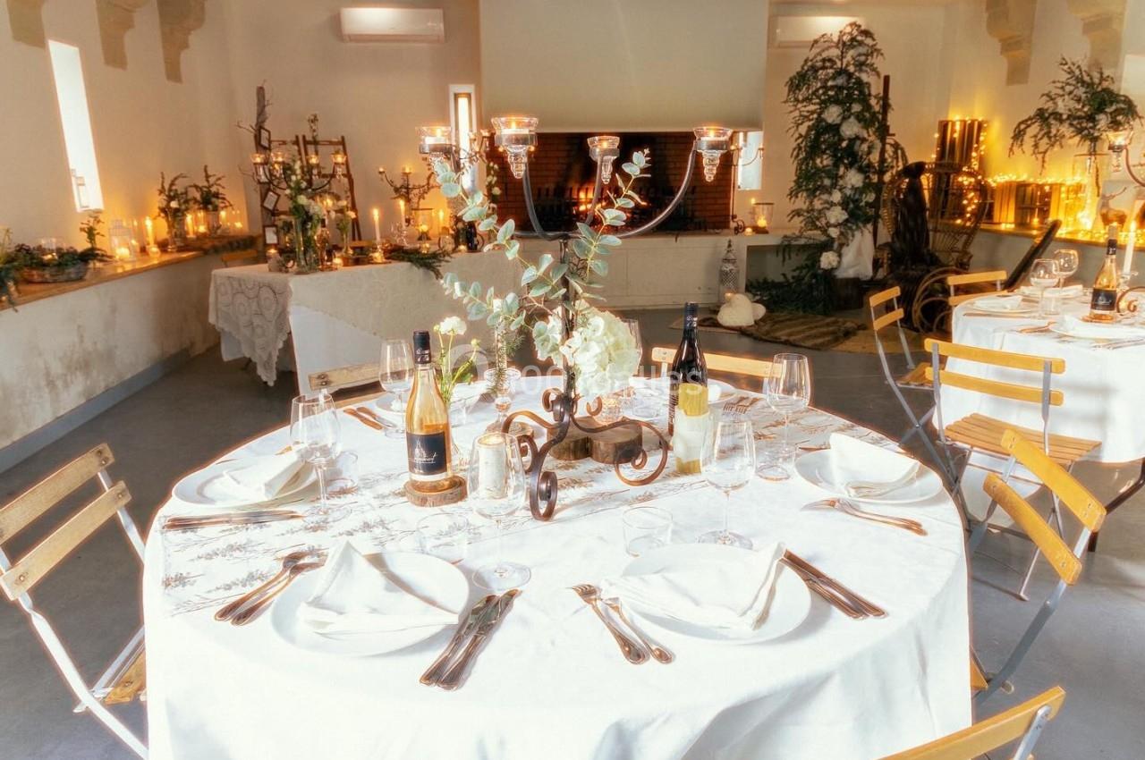 Table dressée avec nappes blanches, couverts, verres et bouteilles de vin, dans une salle décorée avec chandeliers et…