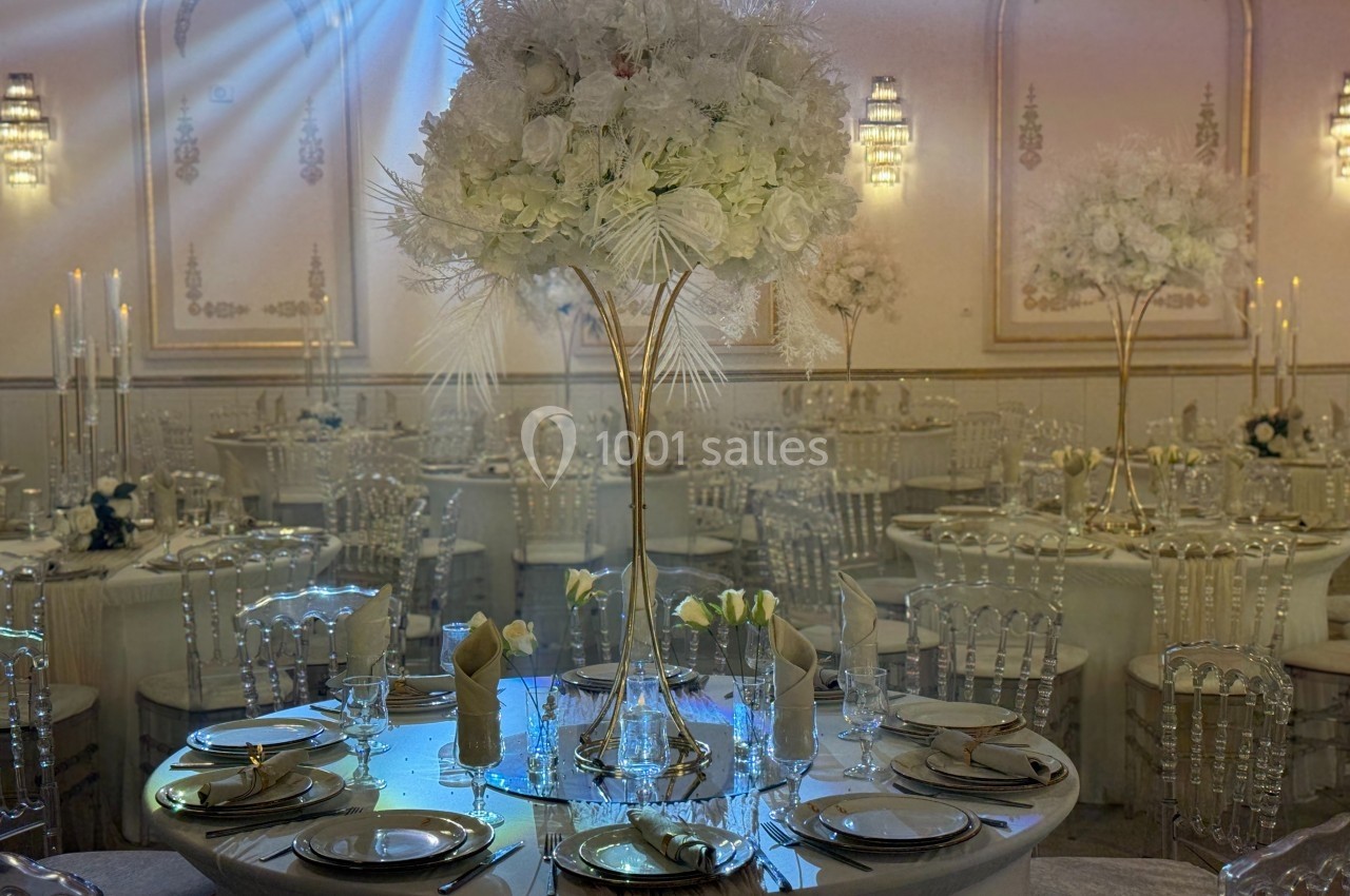 Salle de réception élégante avec tables dressées, centres de table floraux blancs et chaises transparentes.