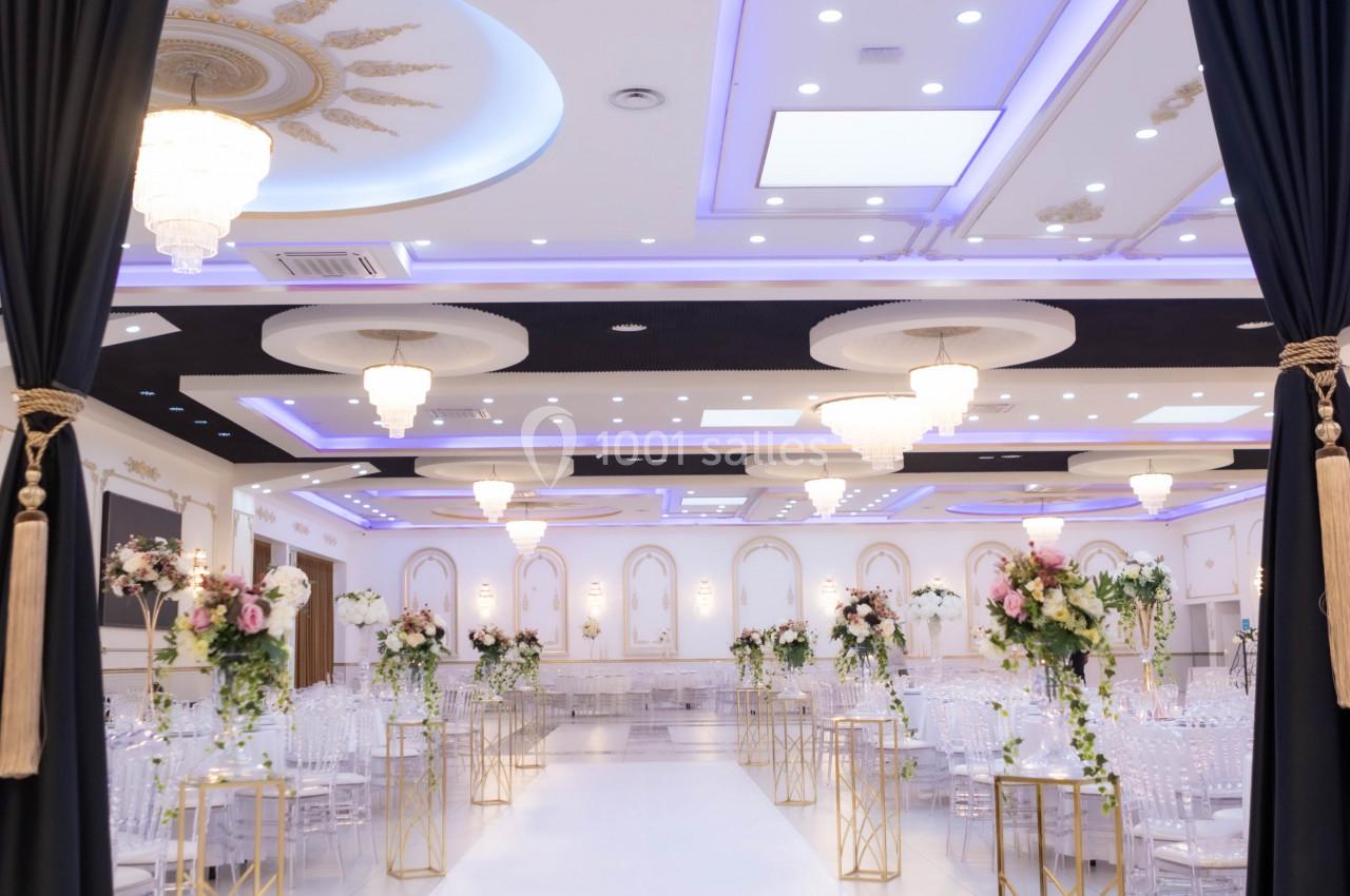 Salle de réception élégante avec allée centrale bordée de fleurs, éclairage lumineux et décorations raffinées.