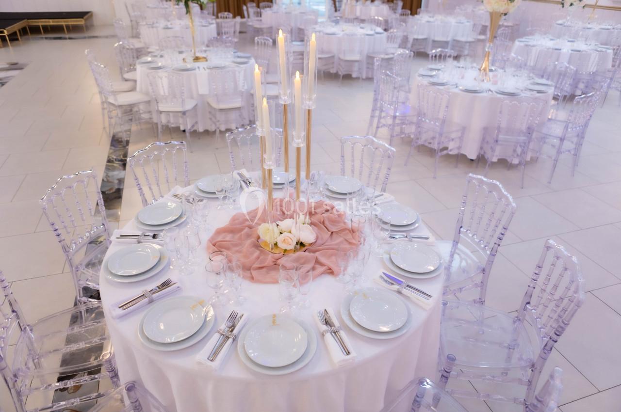 Table ronde élégamment dressée avec nappes blanches, chaises transparentes, centre de table floral et bougies hautes.