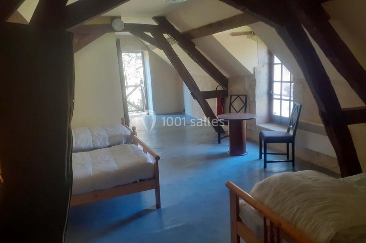 Chambre mansardée avec poutres apparentes, deux lits simples, une table et une chaise près d'une fenêtre.