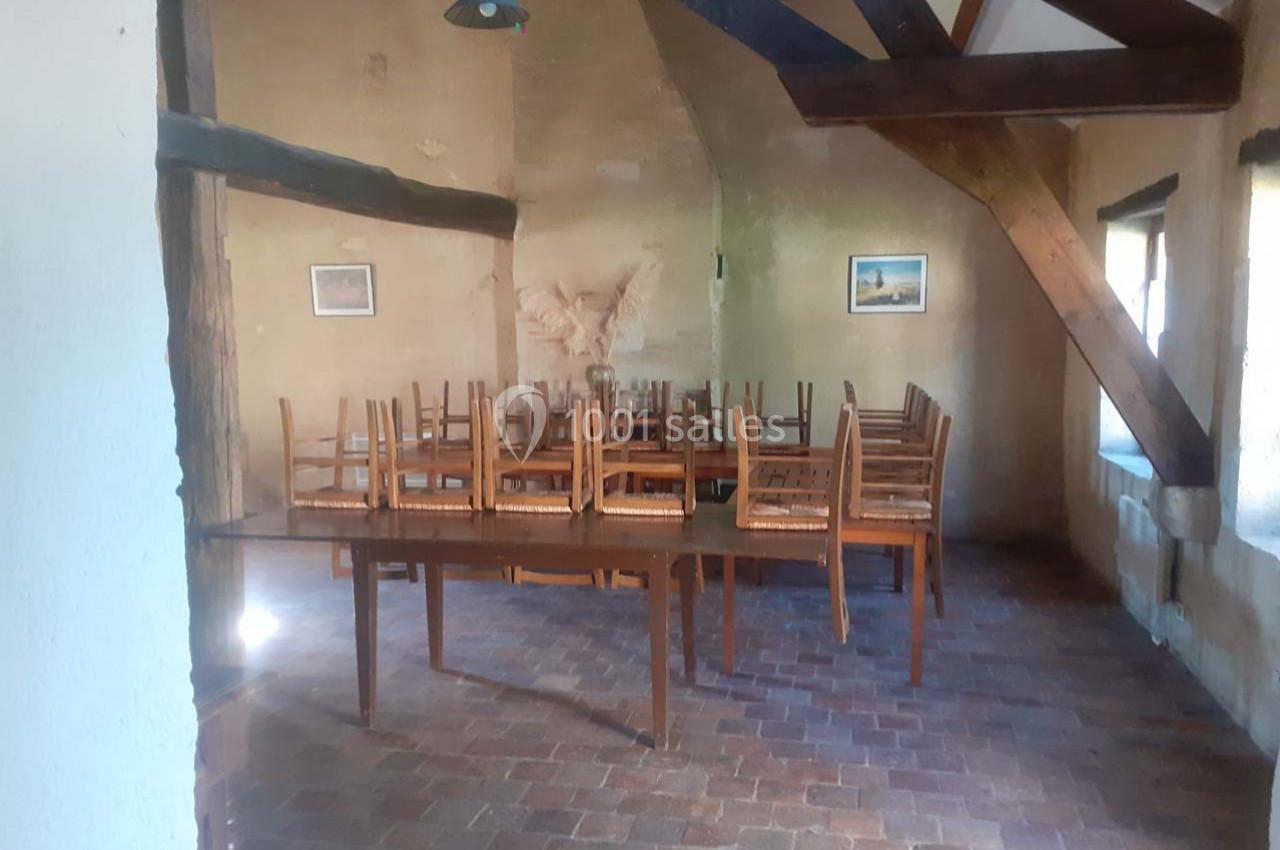 Salle rustique avec poutres apparentes, tables en bois et chaises empilées, éclairée par des fenêtres latérales.