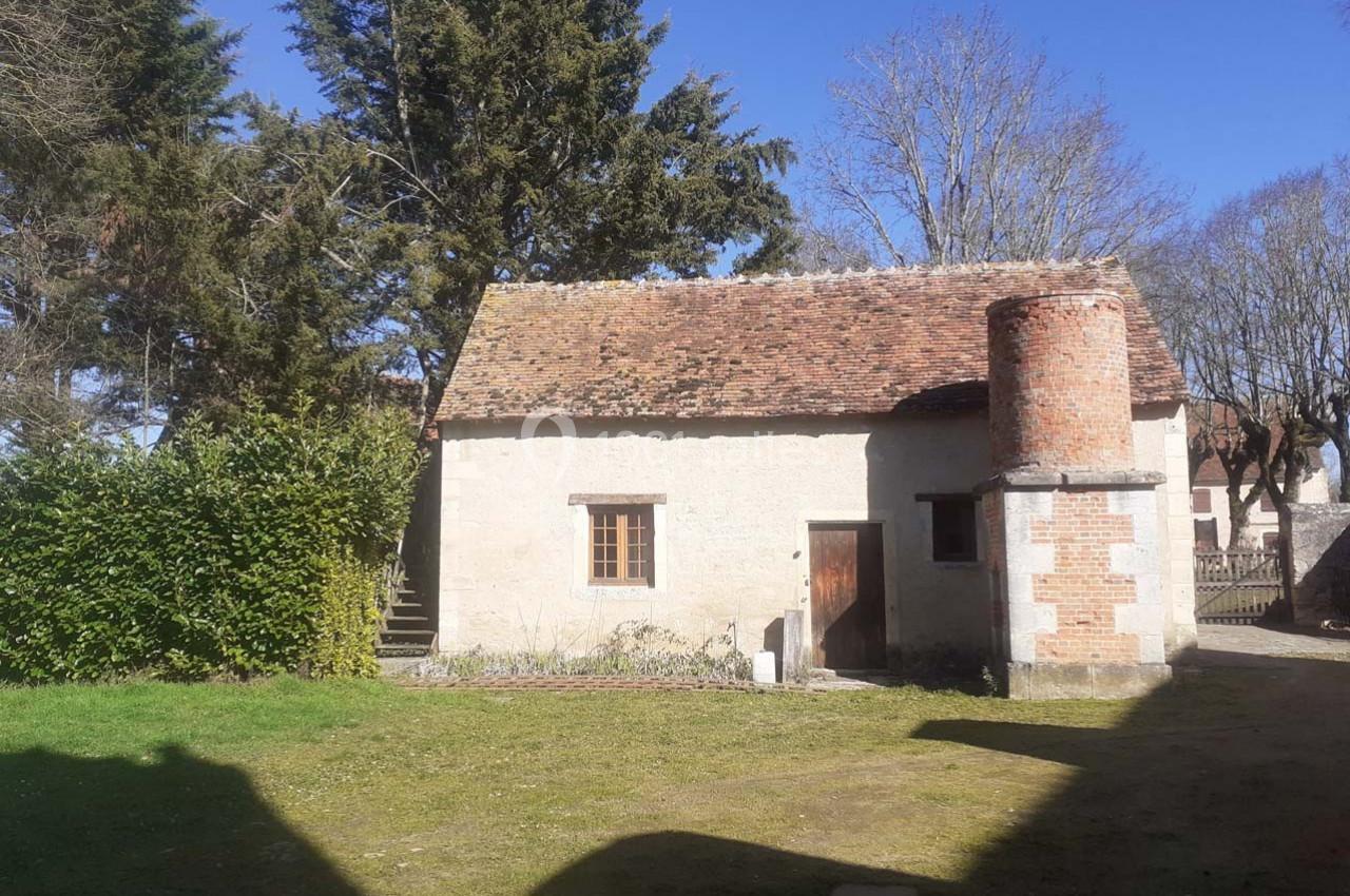 Petite maison en pierre avec toit en tuiles, cheminée en briques, entourée d'arbres et d'une pelouse.