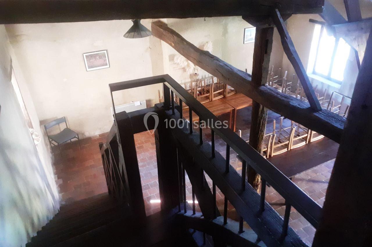 Escalier en bois sombre menant à une salle avec des tables et des chaises, éclairée par une fenêtre.
