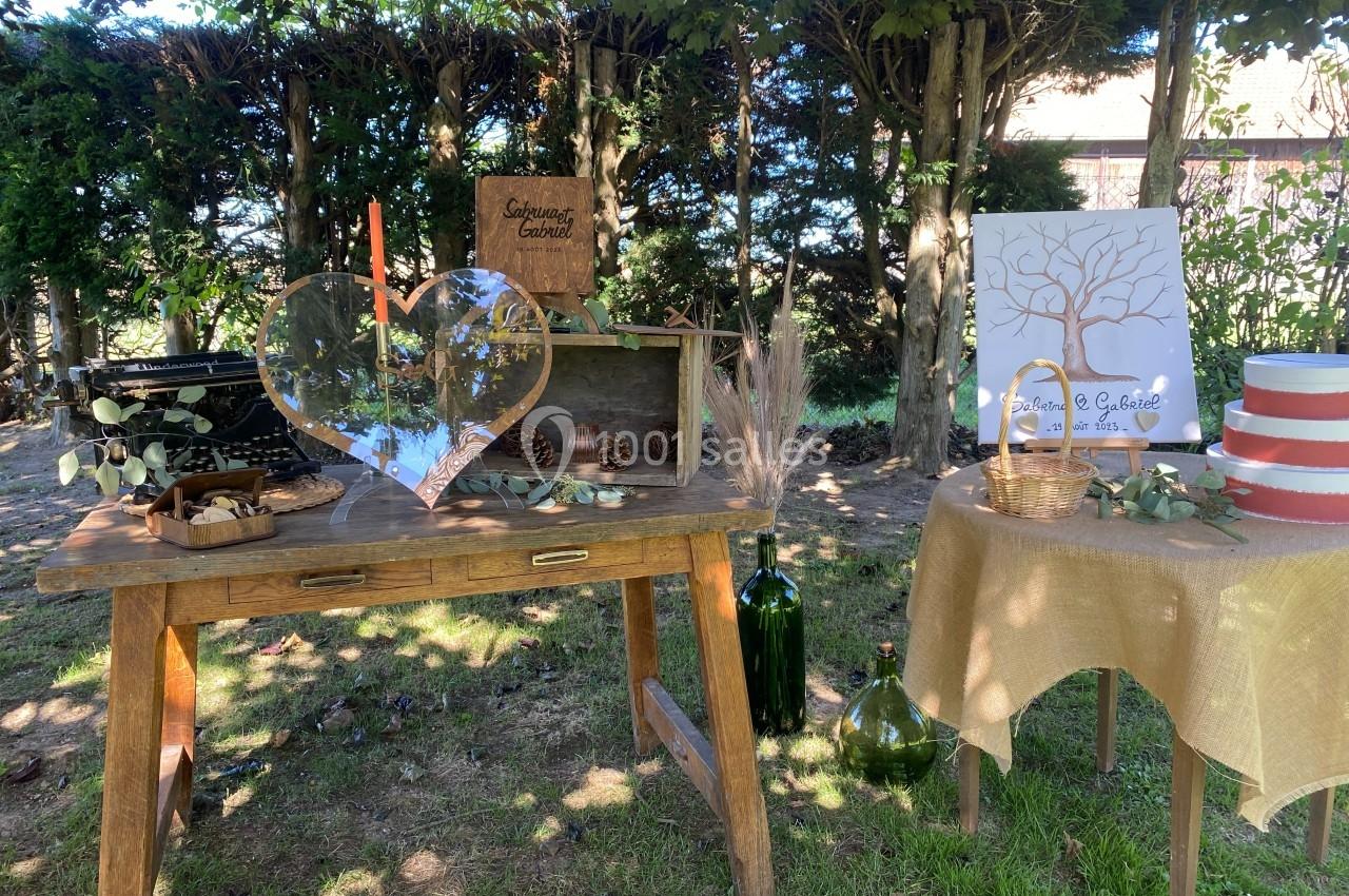 Location salle Beaubray (Eure) - Auberge de la Comtesse #6 Décoration champêtre avec table en bois, objets rustiques, cœur en verre et arbre à empreintes sous des arbres.