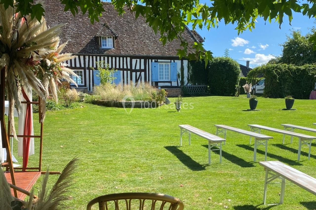 Location salle Beaubray (Eure) - Auberge de la Comtesse #12 Jardin verdoyant avec bancs blancs disposés devant une maison à colombages sous un ciel bleu.