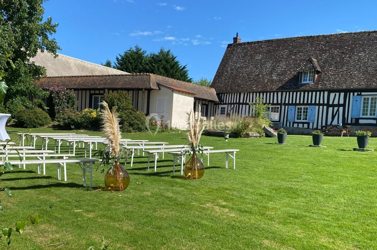 Location salle Beaubray (Eure) - Auberge de la Comtesse #7 Jardin avec pelouse aménagée pour un événement, bancs blancs alignés et décorations florales devant un bâtiment à colombages.
