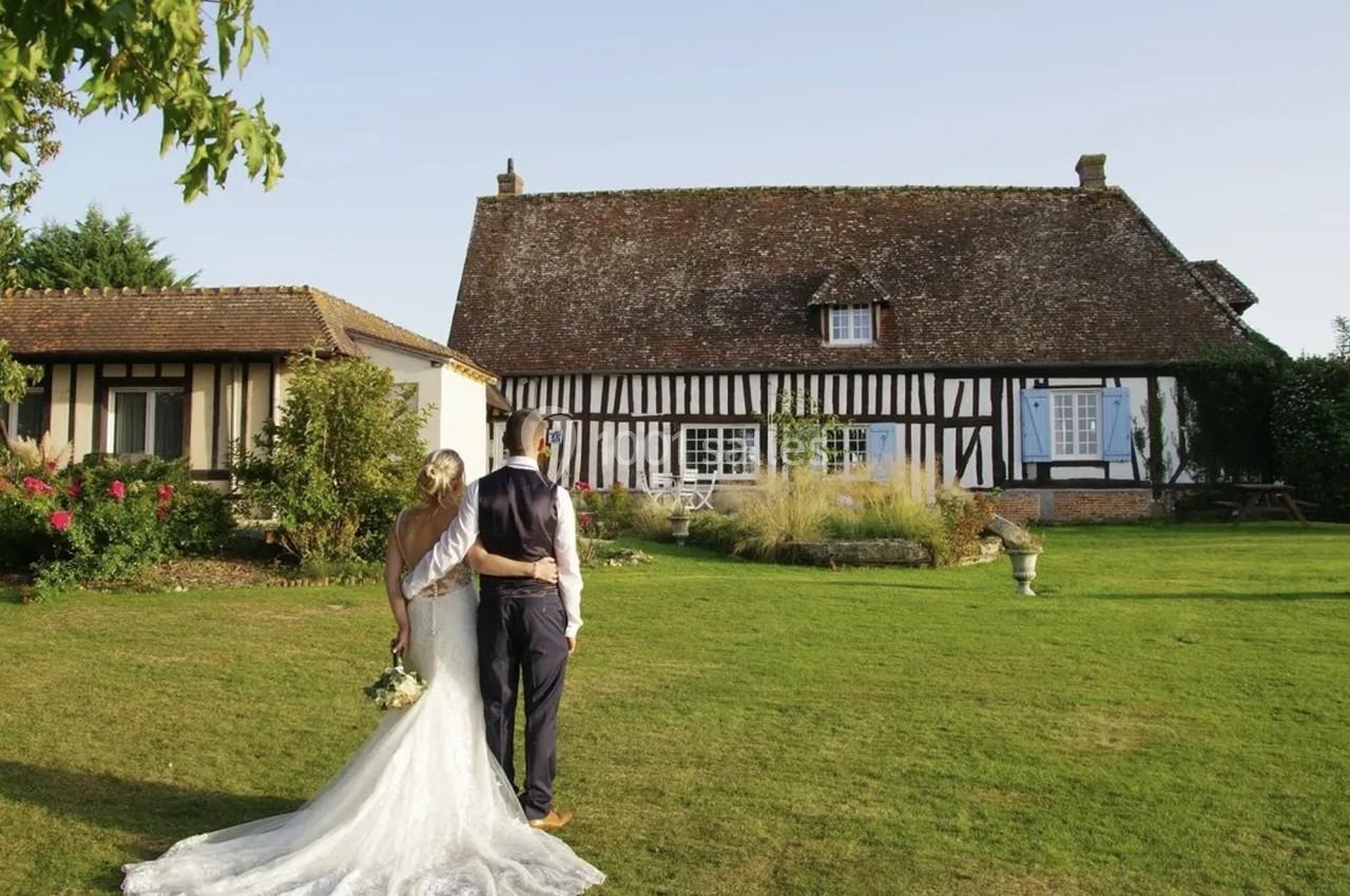 Location salle Beaubray (Eure) - Auberge de la Comtesse #4 Un couple en tenue de mariage regarde une maison à colombages dans un jardin verdoyant par une journée ensoleillée.