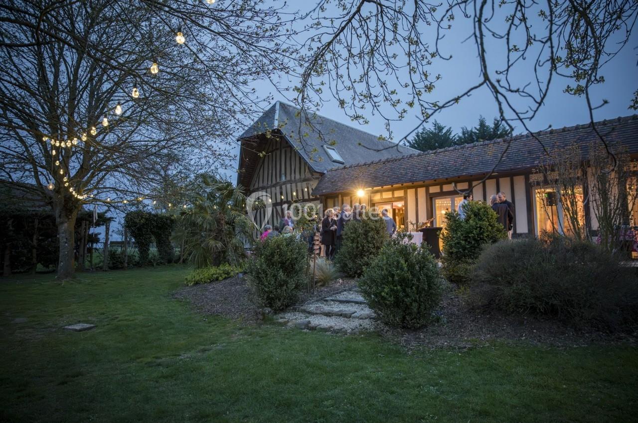 Location salle Beaubray (Eure) - Auberge de la Comtesse #5 Maison à colombages éclairée en soirée, entourée d'arbres et de pelouse, avec des invités rassemblés à l'extérieur.