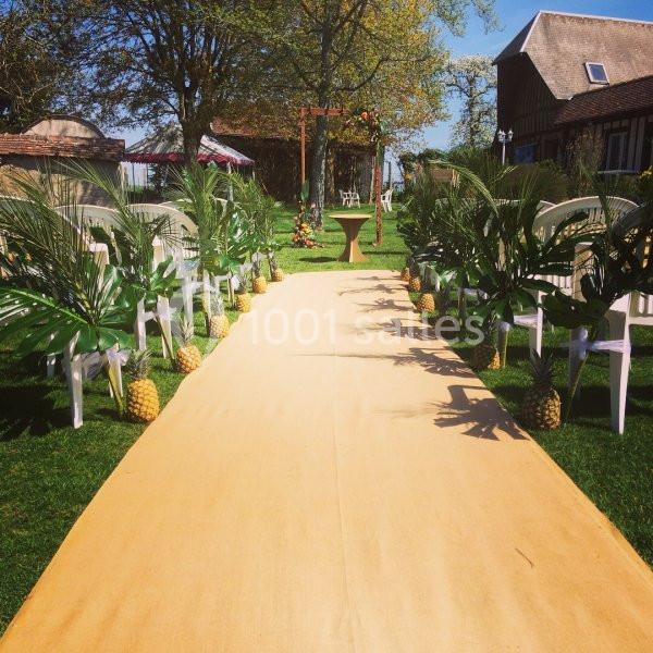Cérémonie laïque - Mariage thème voyage - Auberge de la comtesse Allée de cérémonie extérieure bordée de feuilles tropicales et d'ananas, avec des chaises blanches disposées de chaque côté.