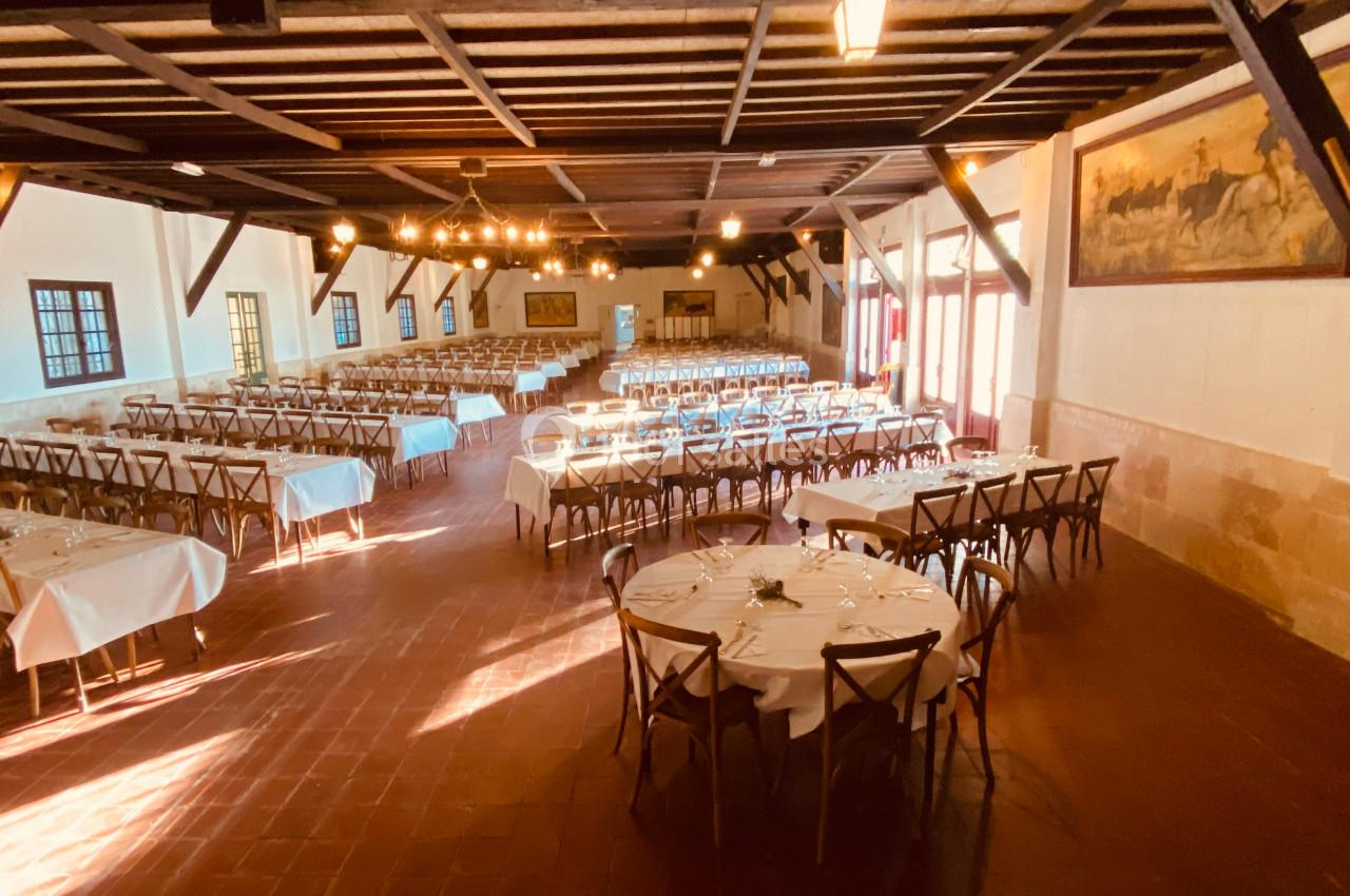 Grande salle de réception avec tables rondes et rectangulaires dressées, éclairée par des lustres et la lumière naturelle.