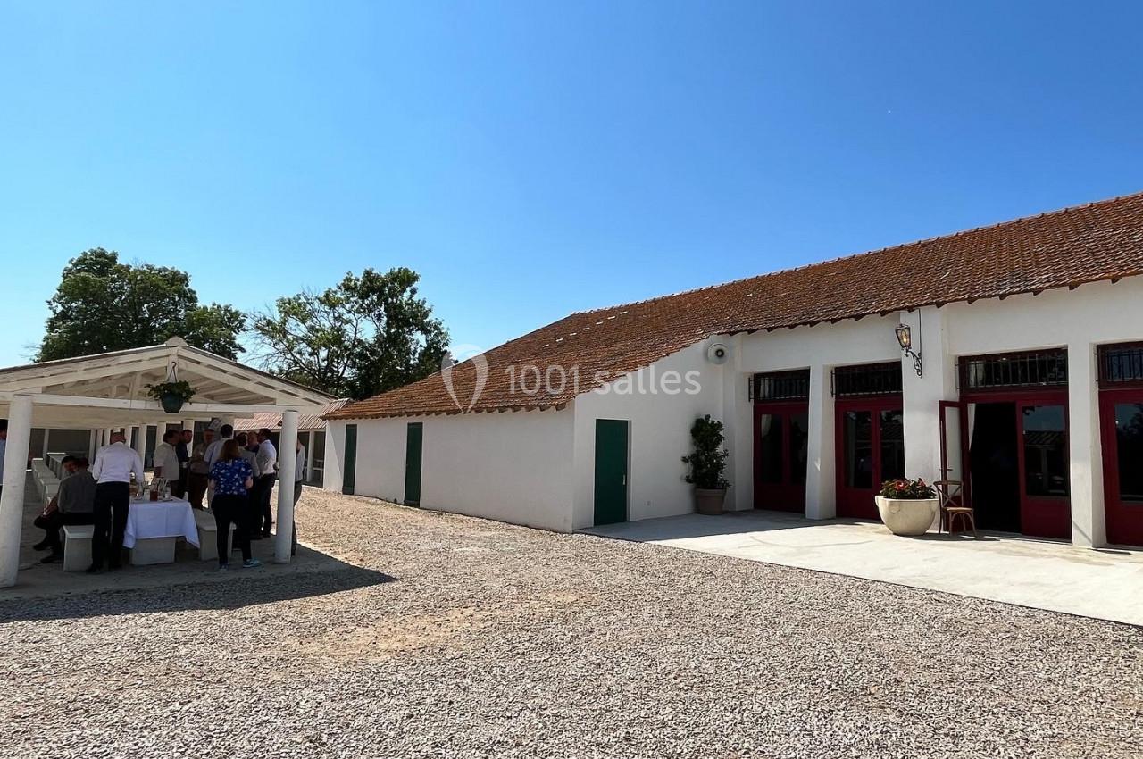 Un groupe de personnes se tient sous un auvent près d'un bâtiment blanc avec des portes rouges, par une journée ensoleillée.