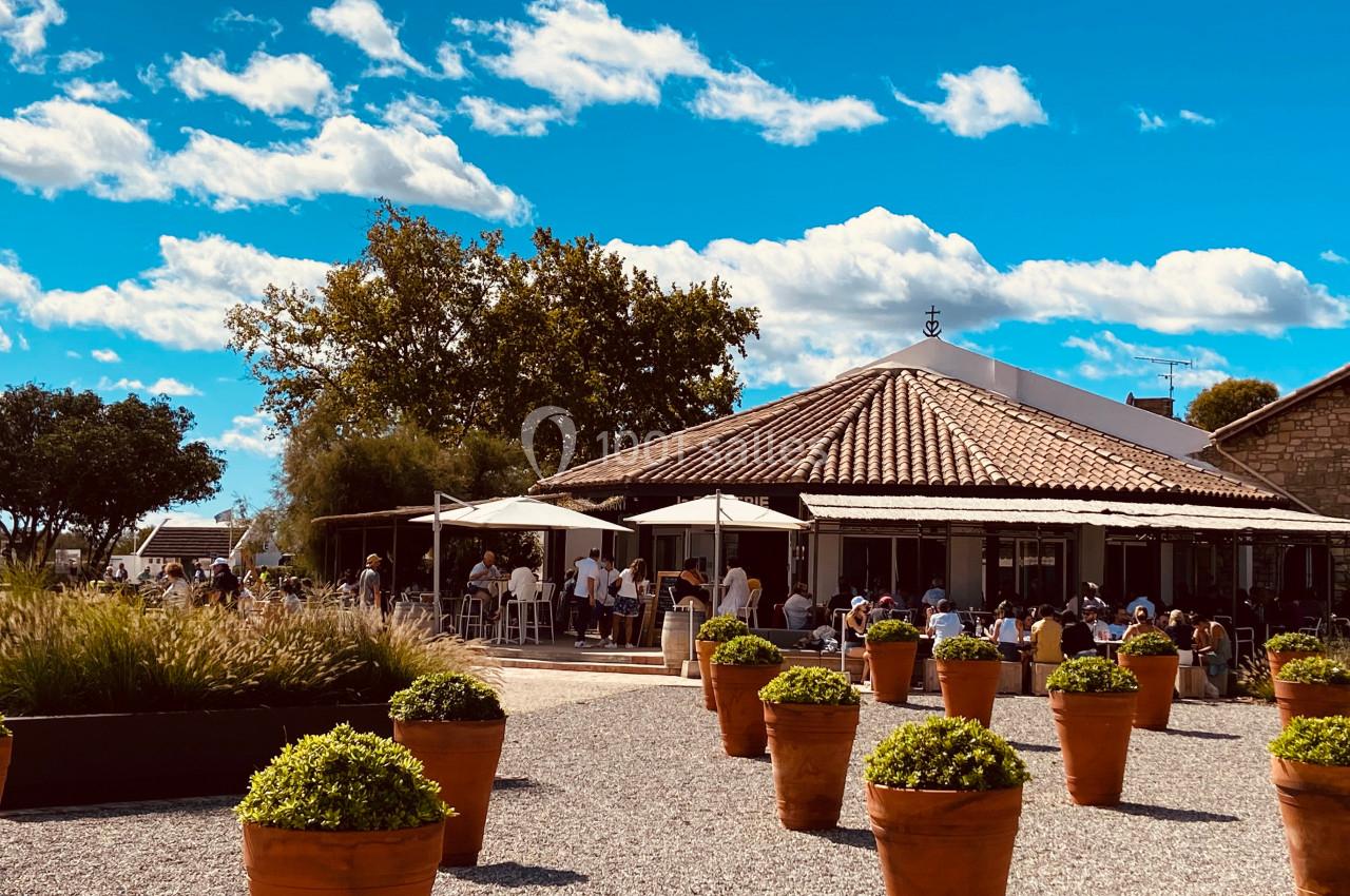 Terrasse d'un restaurant avec des tables ombragées, entourée de pots de plantes, sous un ciel bleu parsemé de nuages.