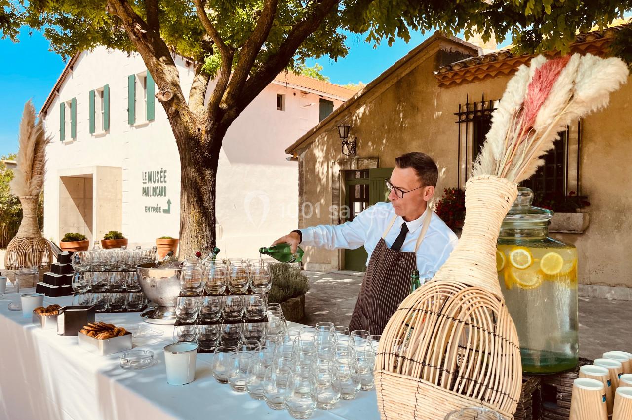 Un serveur verse une boisson dans un verre sur une table dressée en extérieur, près d'un bâtiment provençal.