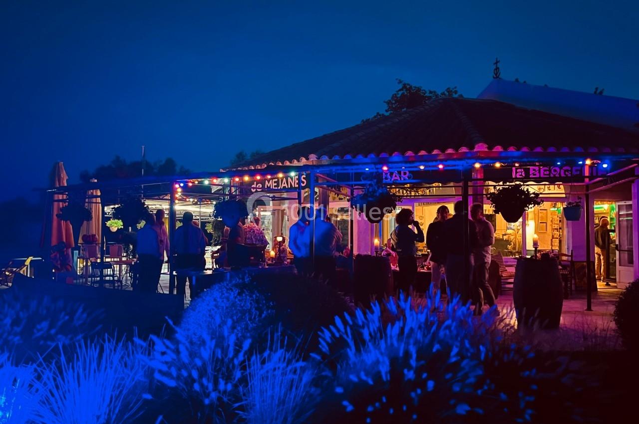 Terrasse d'un bar-restaurant éclairée par des lumières colorées en soirée, avec des clients debout et assis.