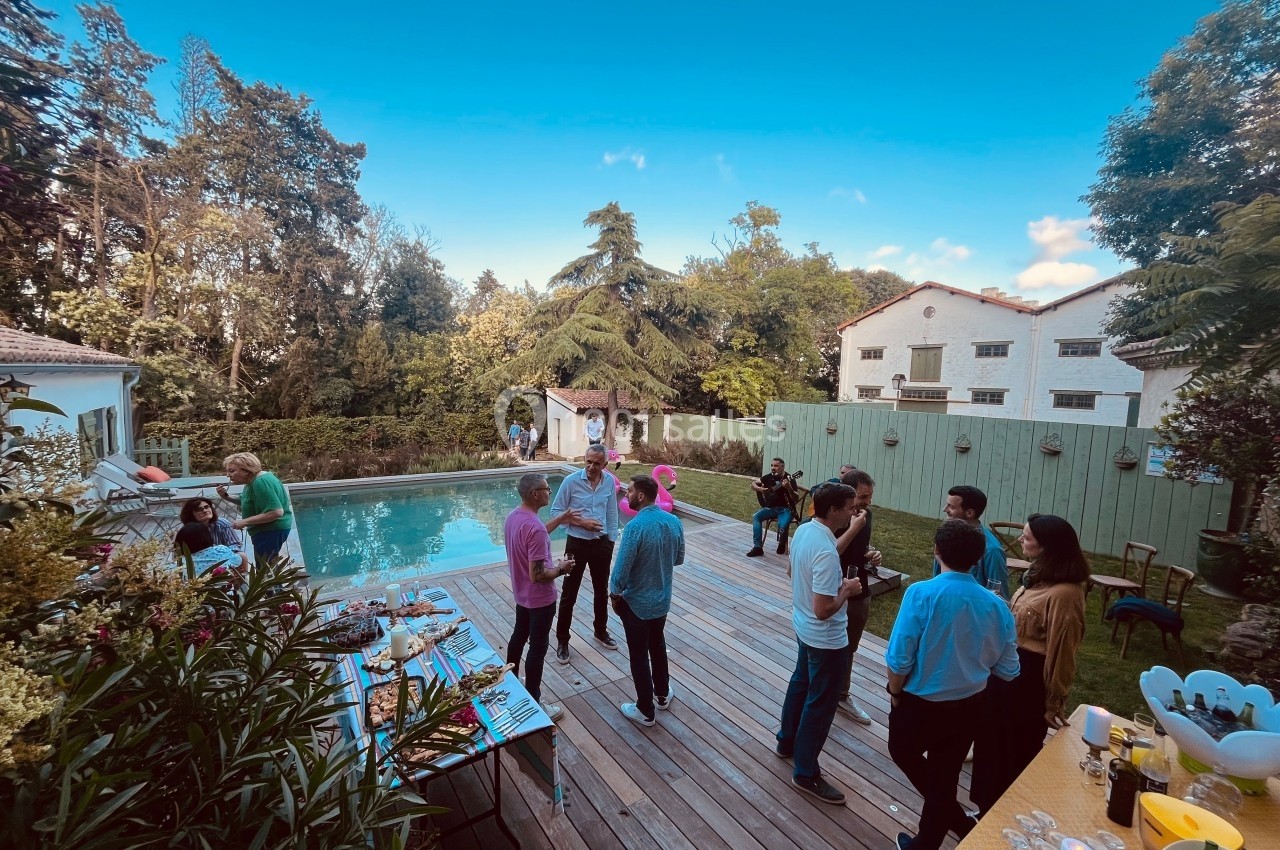 Personnes discutant sur une terrasse en bois près d'une piscine, entourée de végétation et de bâtiments blancs.