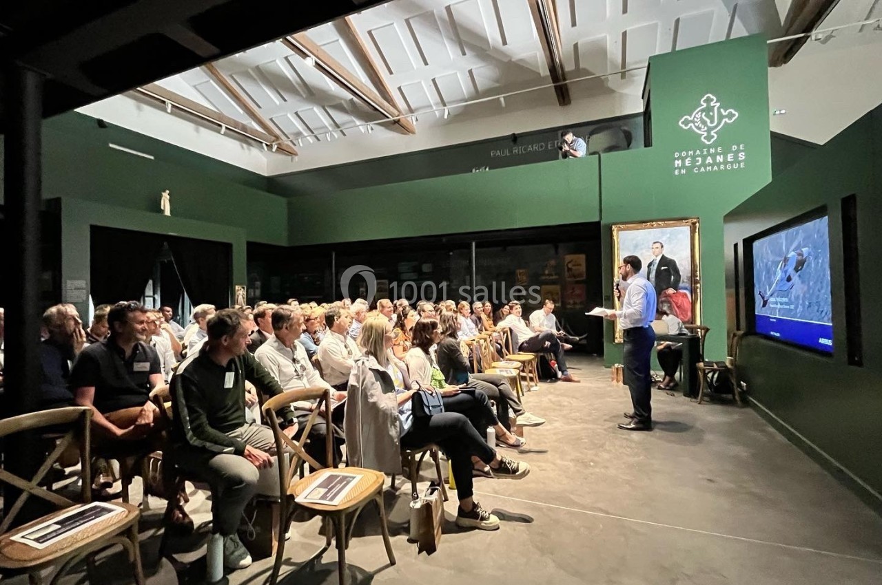 Un conférencier s'adresse à un public assis dans une salle sombre avec un écran et un tableau en arrière-plan.