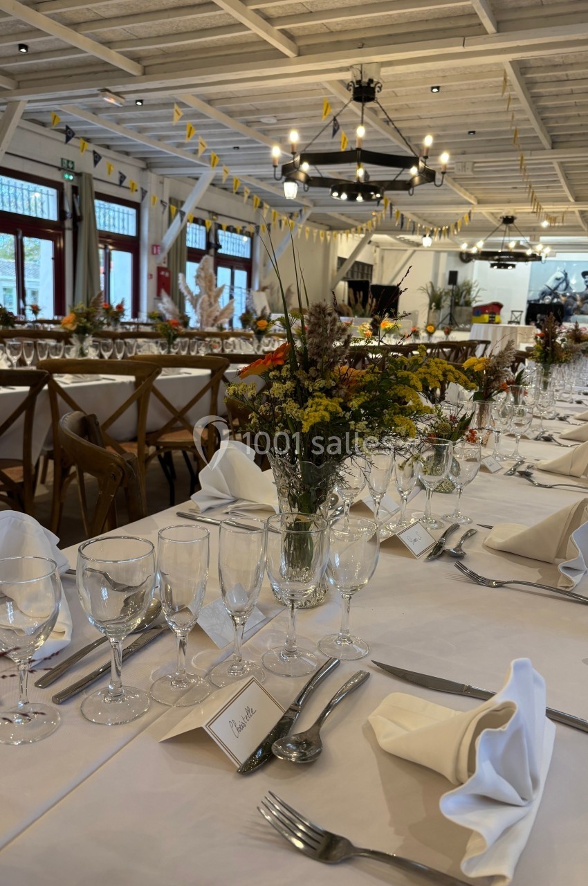 Salle de réception décorée avec des tables dressées, nappes blanches, verres, couverts et bouquets de fleurs champêtres.