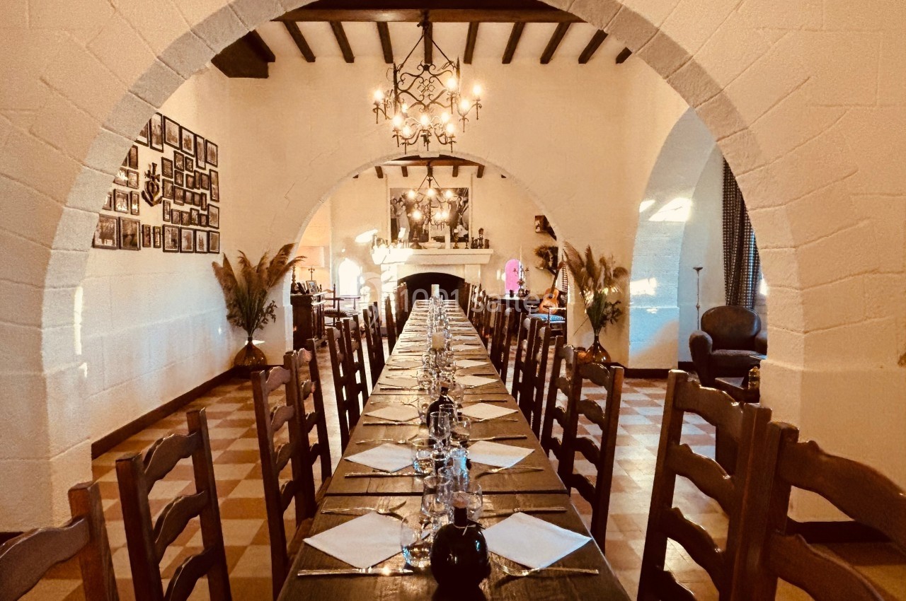 Salle à manger rustique avec une longue table en bois, chaises en bois, poutres apparentes et éclairage chaleureux.