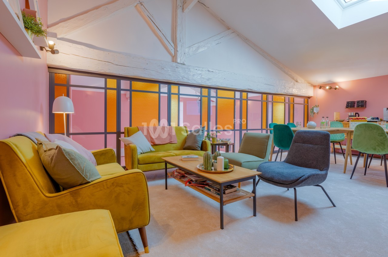 Salon lumineux avec murs roses, mobilier coloré, poutres apparentes et grande verrière laissant passer la lumière naturelle.