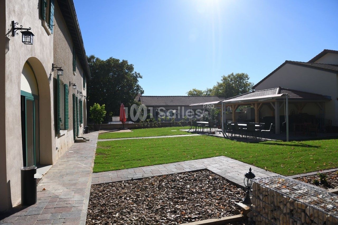 Cour extérieure avec pelouse, terrasse couverte, parasol rouge et bâtiments en arrière-plan sous un ciel ensoleillé.