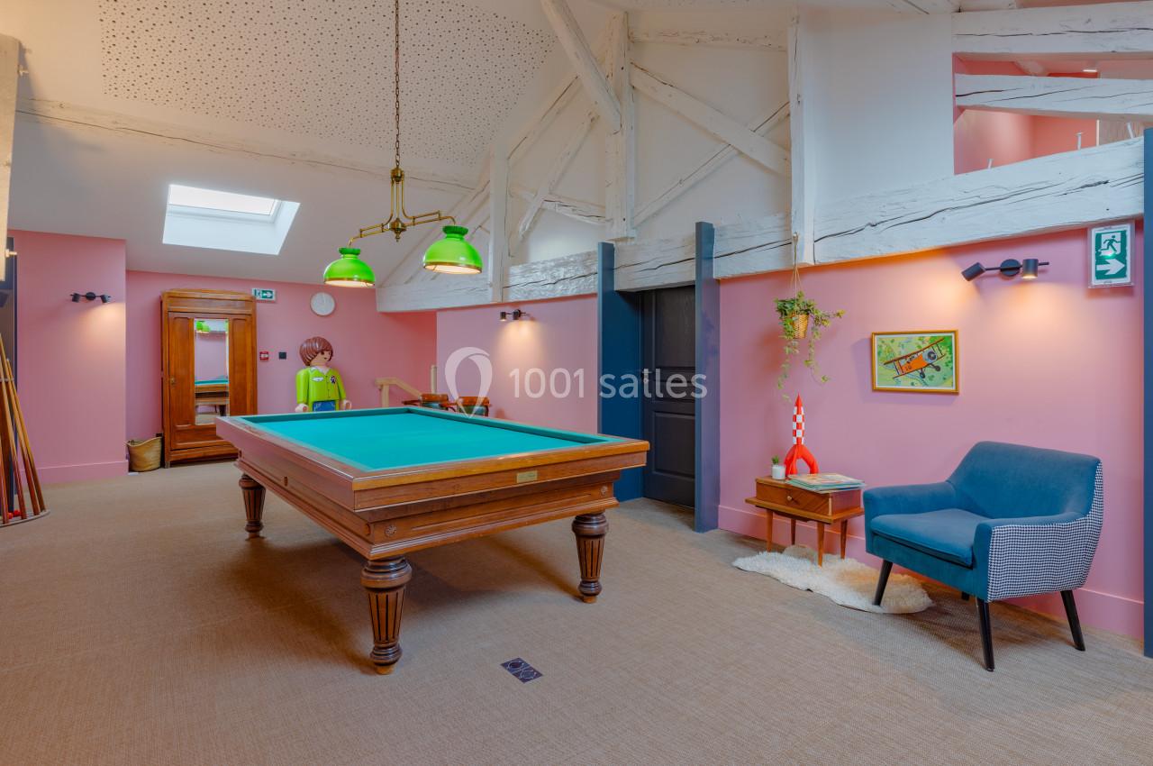 Salle de jeux avec une table de billard, fauteuil bleu, décoration murale et poutres apparentes sous un plafond mansardé.