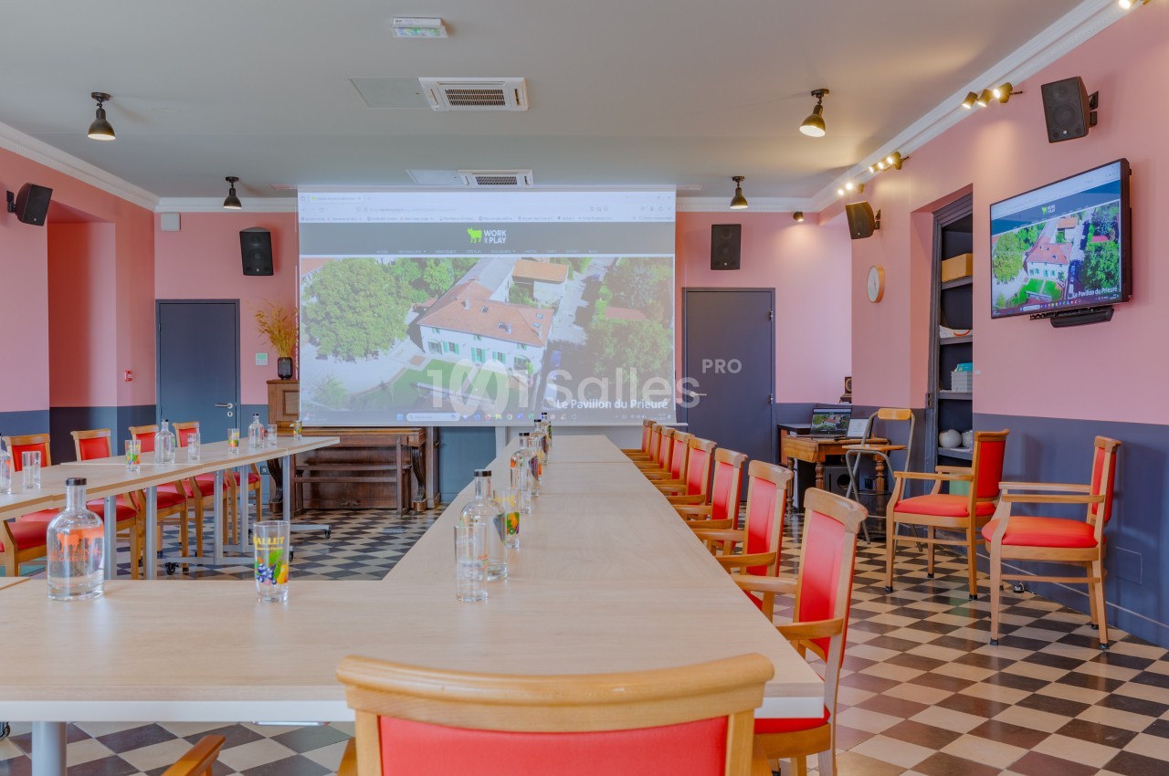 Salle de réunion lumineuse avec tables en U, chaises rouges, écran de projection et décor aux tons roses et bleus.