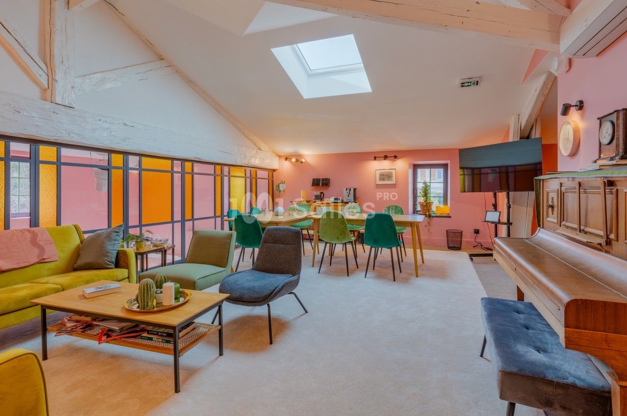 Salle de séjour lumineuse avec murs roses, mobilier coloré, piano, table à manger et grande fenêtre.