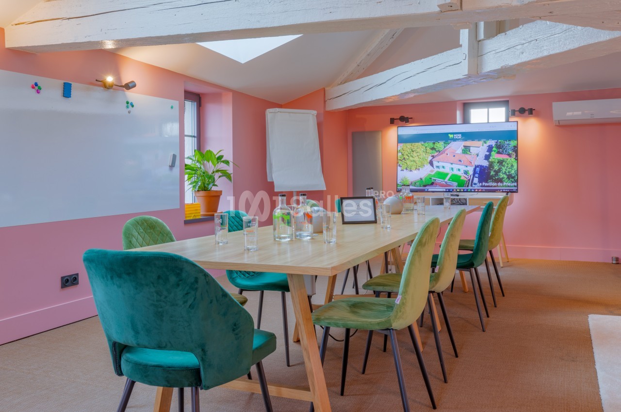 Salle de réunion lumineuse avec table en bois, chaises vertes, écran, tableau blanc et poutres apparentes.