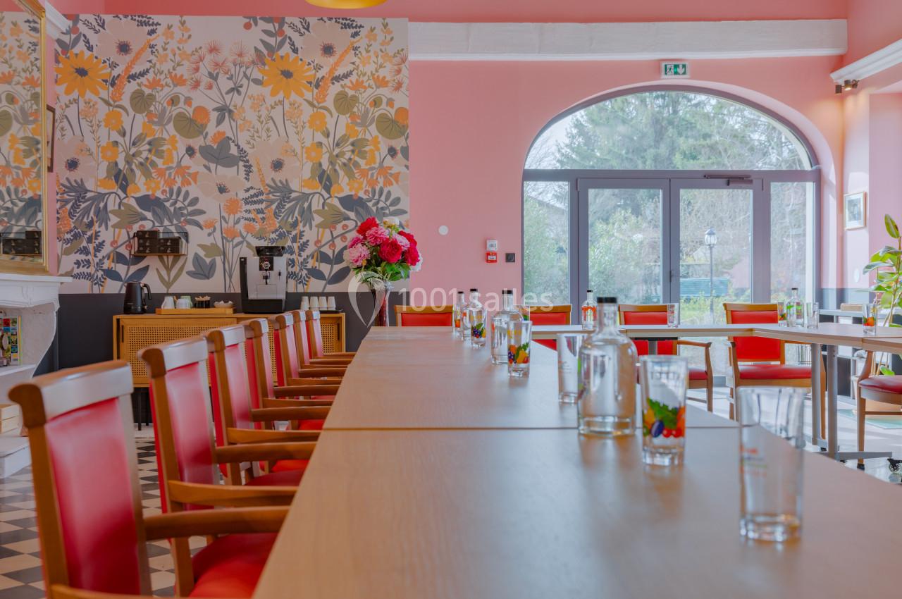 Salle lumineuse avec de longues tables en bois, chaises rouges, mur fleuri et grandes fenêtres donnant sur un jardin.