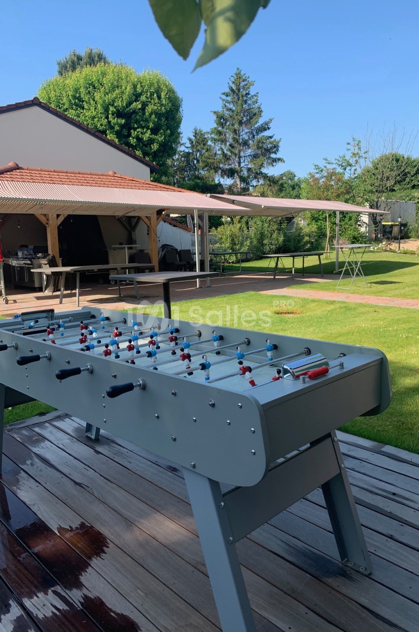Table de baby-foot en extérieur sur une terrasse en bois, avec un jardin et une pergola en arrière-plan.