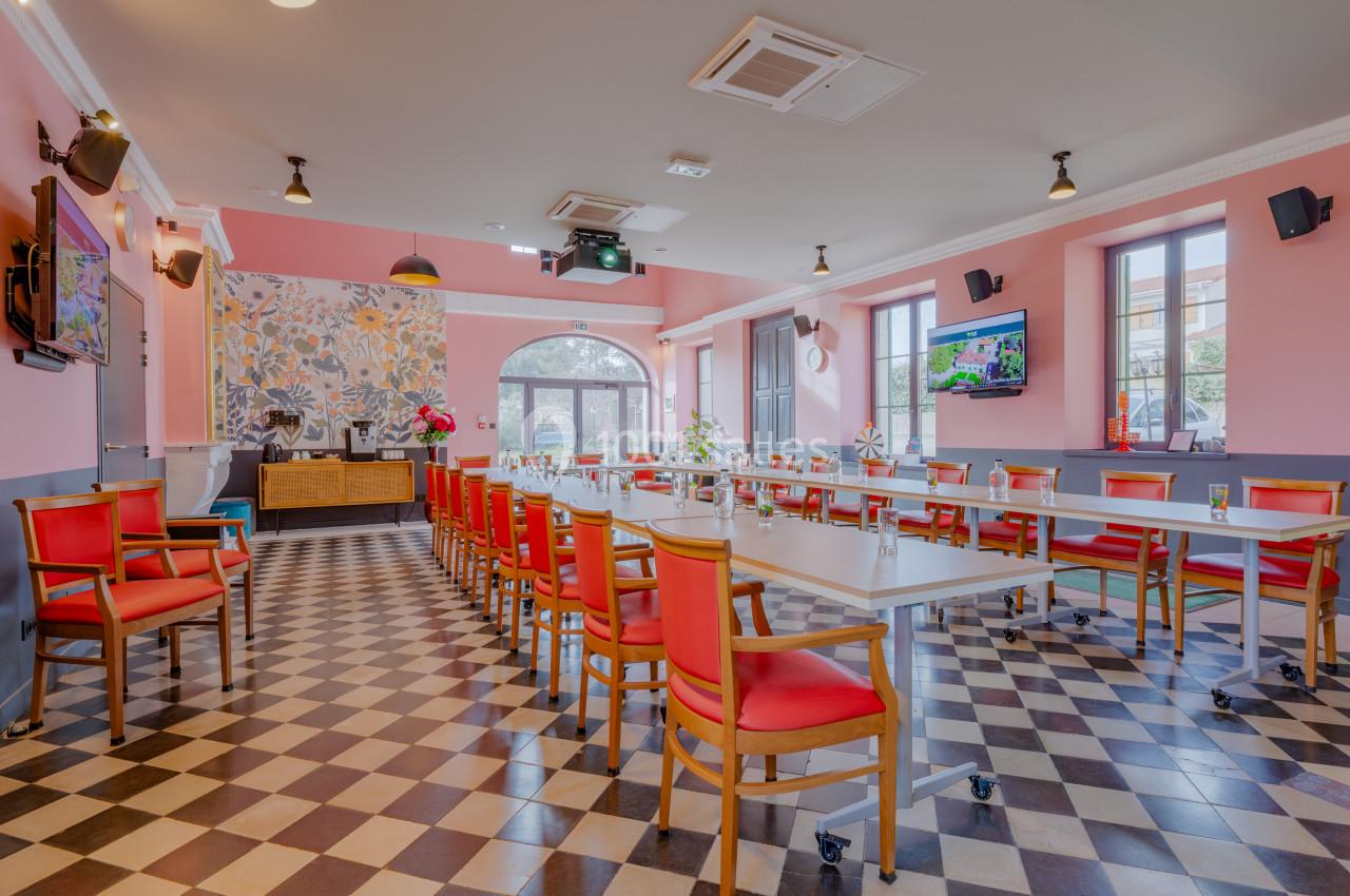 Salle lumineuse aménagée pour une réunion, avec des tables en U, des chaises rouges et un sol à carreaux noir et blanc.