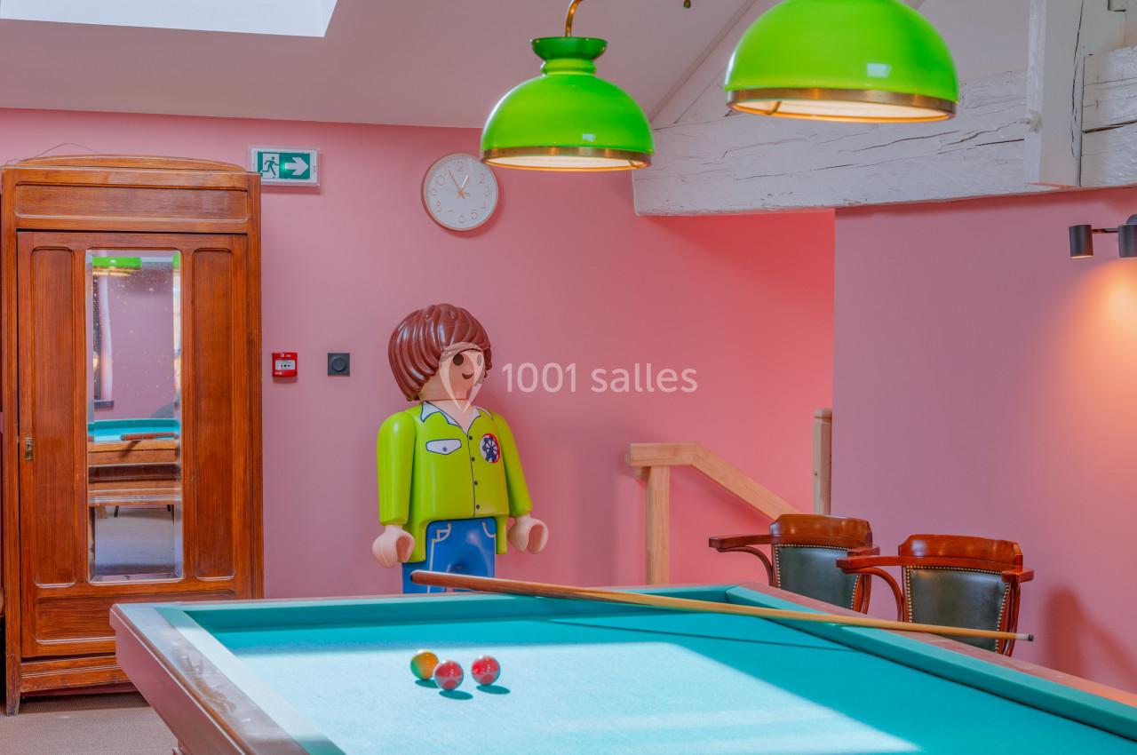 Salle de jeux avec un billard, des lampes vertes suspendues et une grande figurine décorative en arrière-plan.