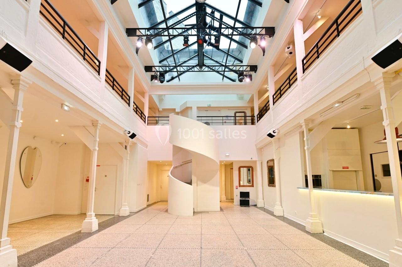 Intérieur lumineux d'un bâtiment avec verrière, escalier en colimaçon central et colonnes blanches.