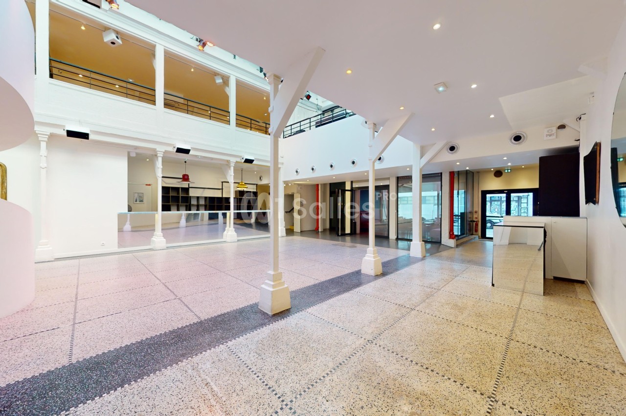 Intérieur d'un espace lumineux avec sol en terrazzo, colonnes blanches, mezzanine et grandes baies vitrées.