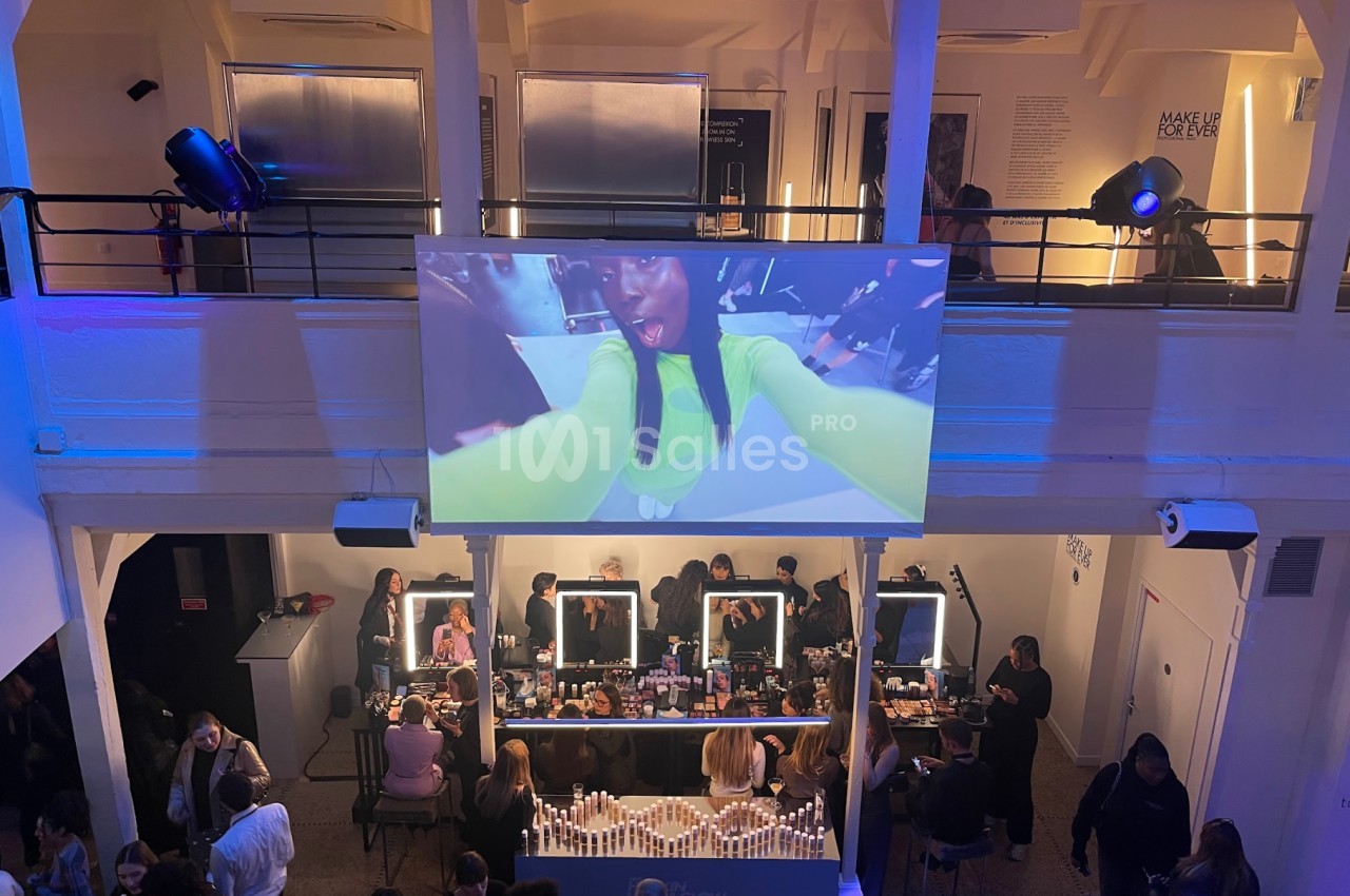 Vue d'un événement avec un écran géant affichant une femme en gros plan et des participants autour de tables de maquillage.